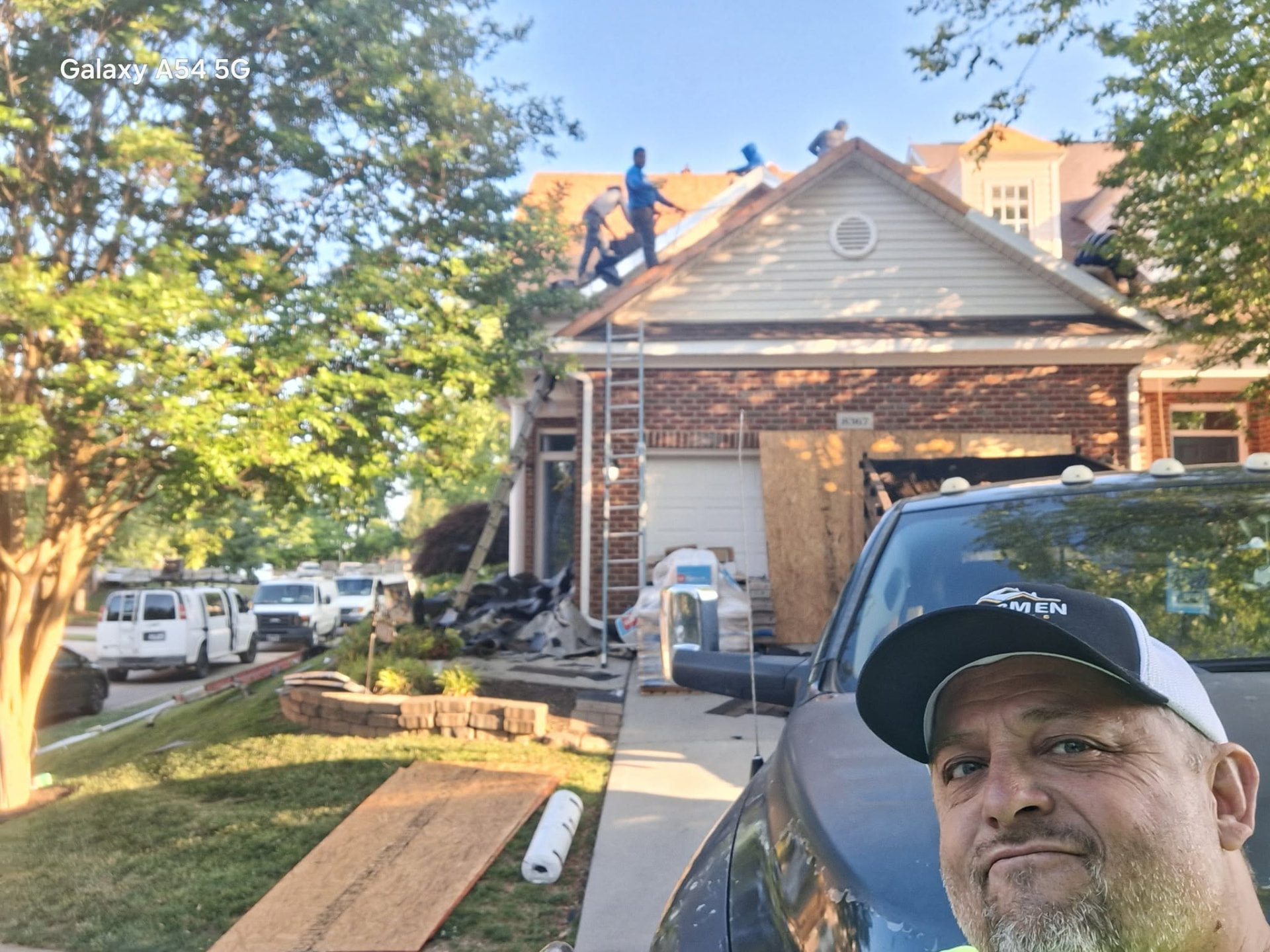 Selfie of a team member at a job site with crew working on a house roof behind him